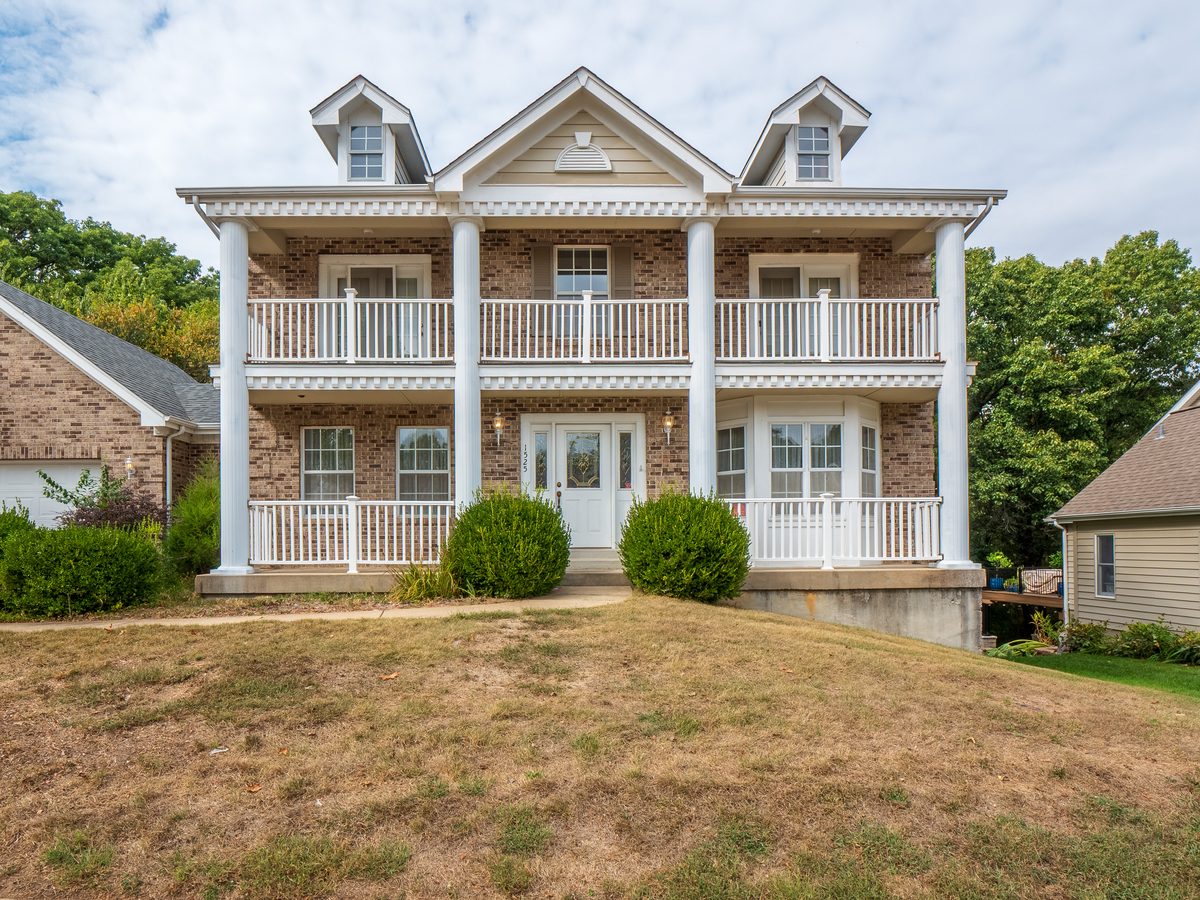 Chesterfield MO homes for sale — 2-story brick colonial with full-width covered porch, upper balcony, and white columns, representative of the larger newer construction buyers find in 63005 west St. Louis County